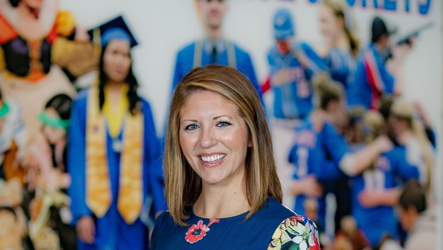A smiling woman in a navy floral dress standing in front of a school banner.