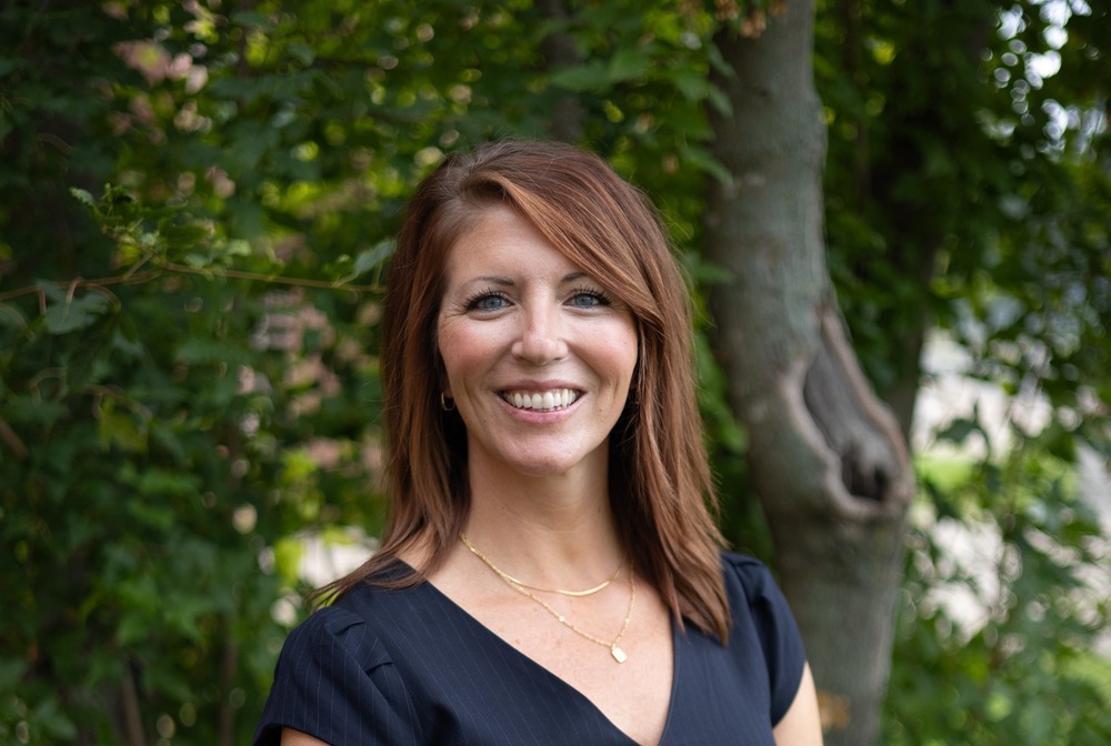 Christina Thayer Anderson smiles in a navy blue dress stands outdoors in front of a tree.