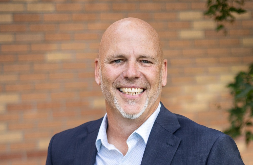 Superintendent Nate Rudolph stands outdoors in front of a brick wall wearing a blue blazer and smiling