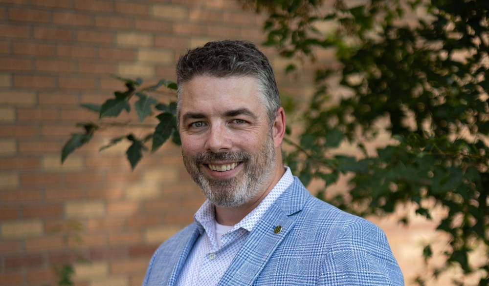 Chris Kampa standing outside in a light blue patterned blazer and white collared shirt, smiling.