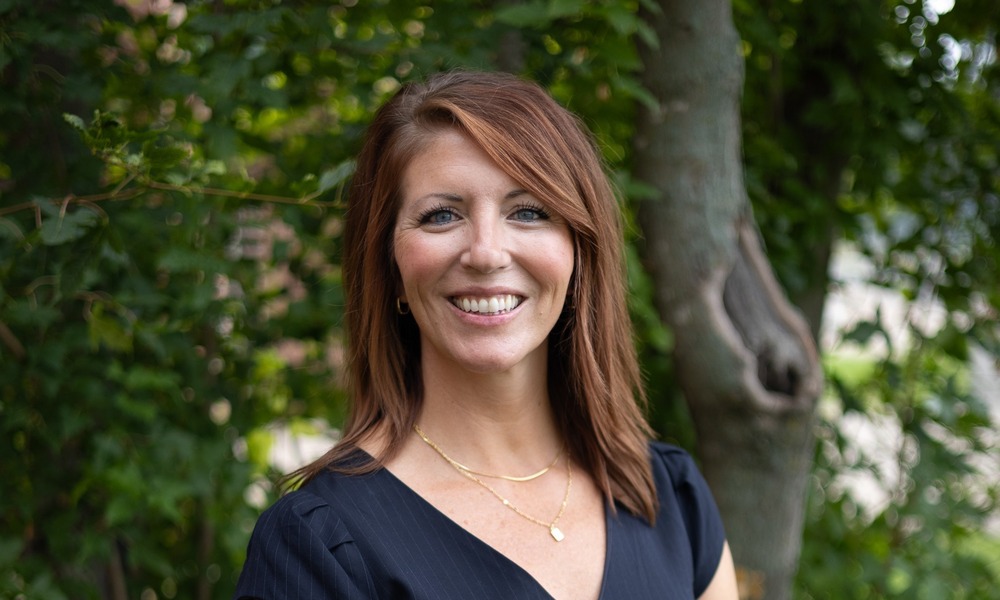 A smiling woman with dark hair stands outdoors against a background of green foliage wearing a navy blue dress.