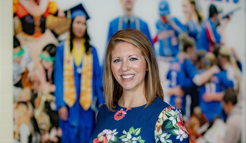 Woman smiling in a floral blue dress, standing in a school hallway with a Blue Jackets wall graphic behind her.