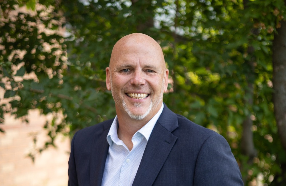 Dr. Nate Rudolph in a dark suit jacket and open-collar shirt standing outdoors in front of trees.