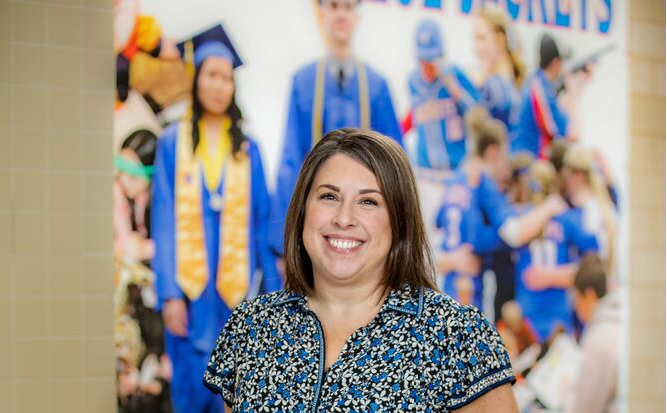 Smiling female staff member standing indoors in a blue and white patterned dress with a school mural in the background.