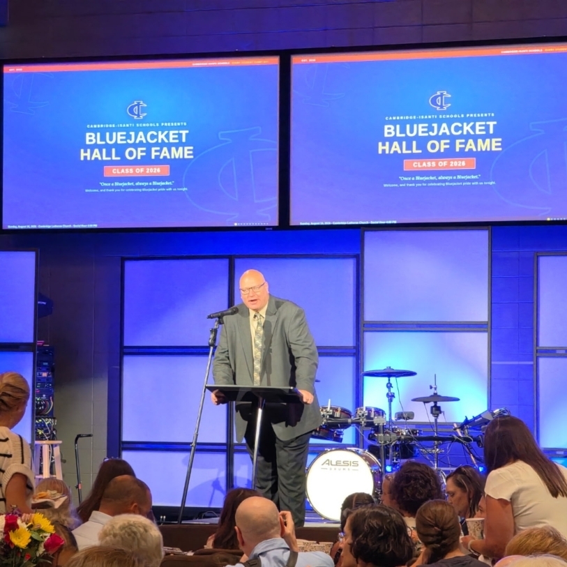 A speaker at a podium addresses a crowd at the Cambridge-Isanti Schools Bluejacket Hall of Fame Class of 2026 event.
