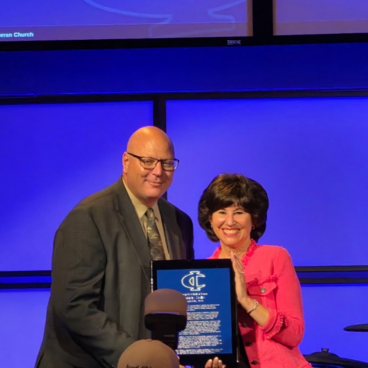 Two people present an award plaque on stage at the Cambridge-Isanti Schools Bluejacket Hall of Fame event.
