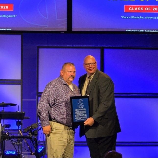 A speaker presents an award plaque to a recipient on stage at the Bluejacket Hall of Fame event.

