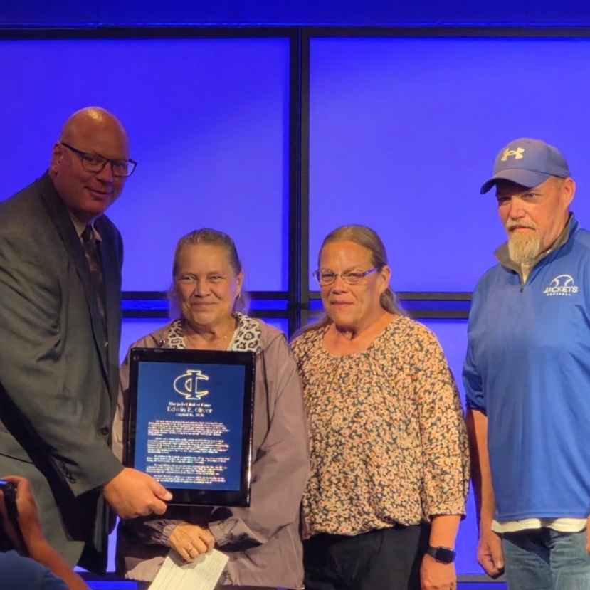 A speaker presents a Bluejacket Hall of Fame award plaque for Edwin R. Oliver to a family on stage at an event.
