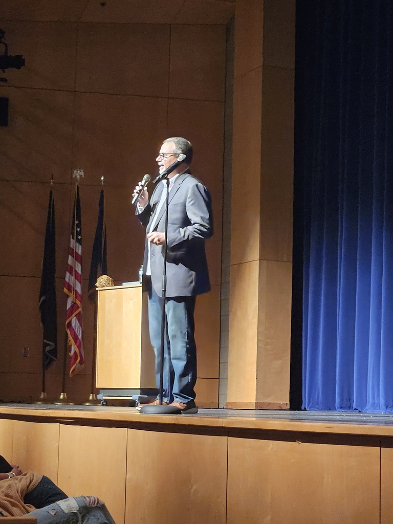 A speaker in a suit addresses an audience from a stage, holding a microphone next to a podium and flags.