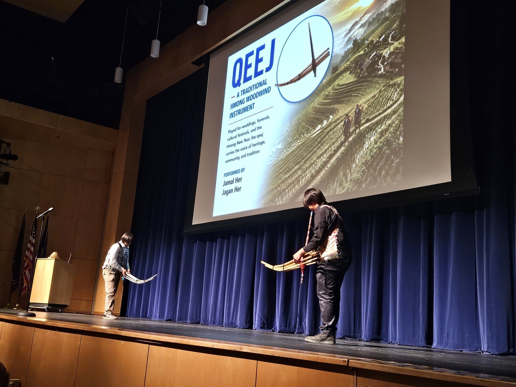 Two musicians perform on a stage in front of a projection screen titled Qeej - A Traditional Hmong Woodwind Instrument.