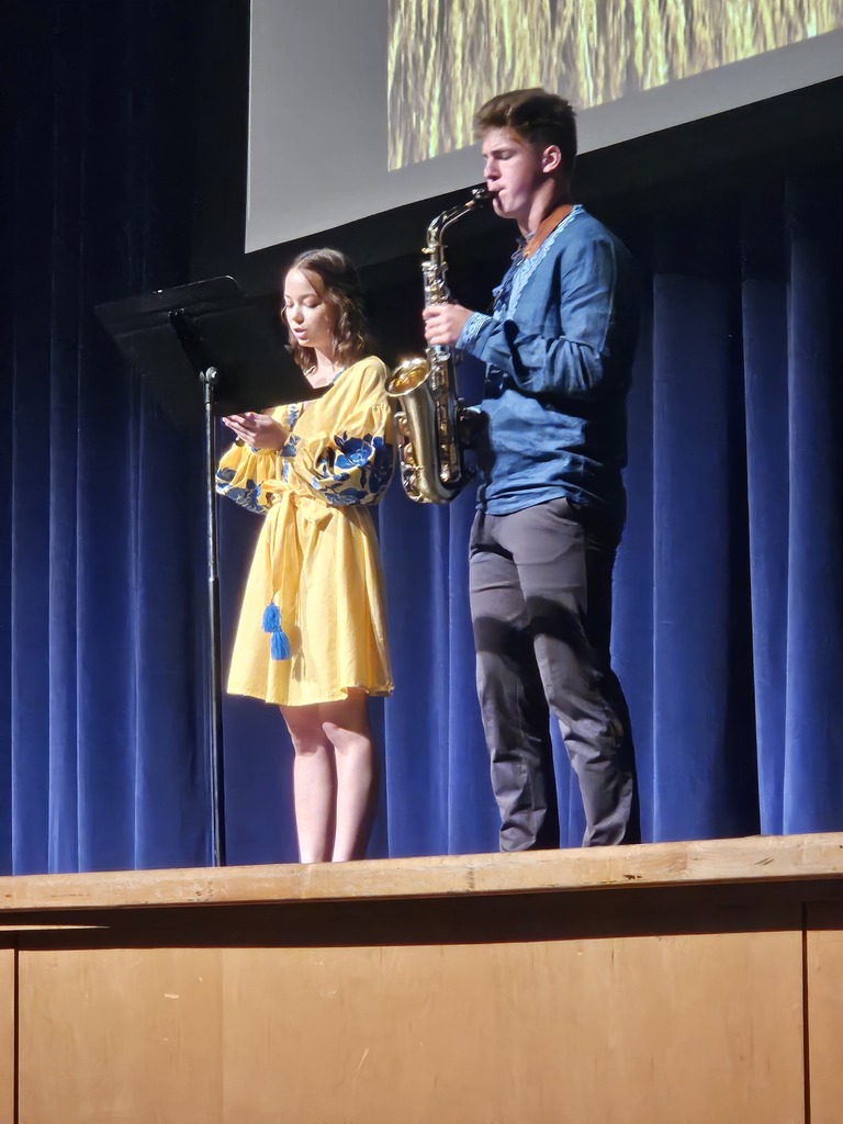 A female student sings into a music stand next to a male student playing the saxophone on stage during a performance.