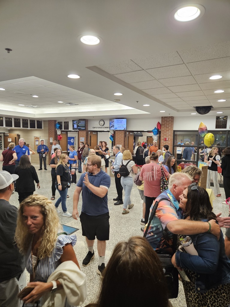 People gather in a crowded school hallway and commons area, talking and greeting each other during a school event.