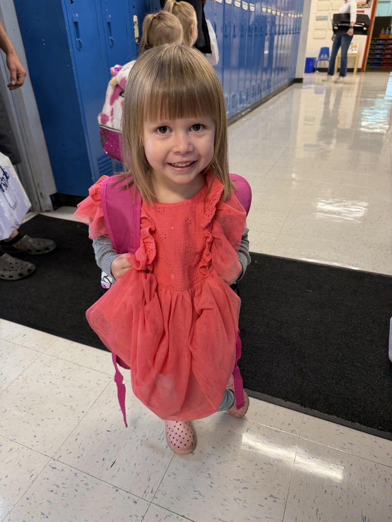 Young girl in a pink dress, wearing a backpack and smiling.