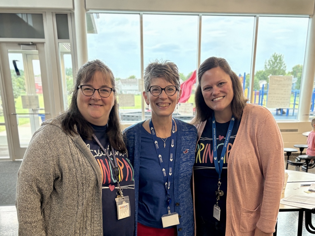 Three school leaders stand together smiling indoors near a window with a playground outside.
