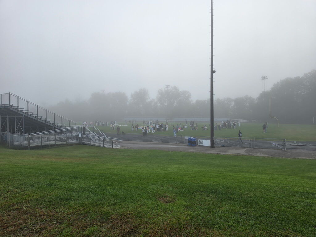 Students gathered on a foggy athletic field, stands to the left.
