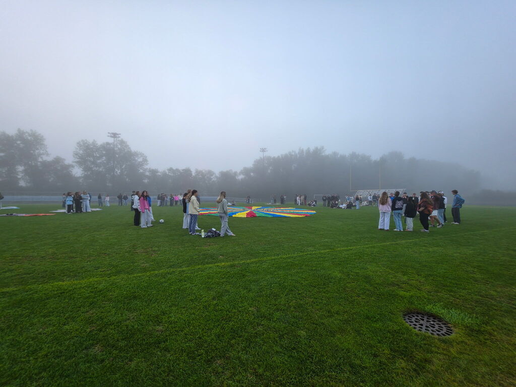 Students gathered in groups on a misty grass field around a large colorful parachute.
