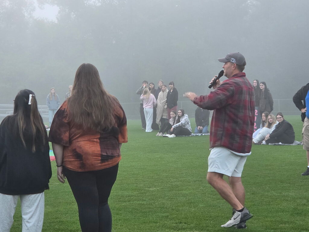 A staff member with a microphone speaks to a group of students gathered on a foggy athletic field.
