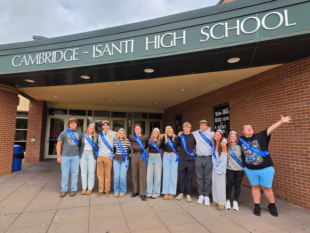 Students wearing sash crowns pose outside Cambridge-Isanti High School in front of a 2026 Homecoming theme sign.
