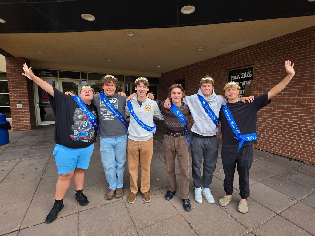 Six male students wearing homecoming sashes and crowns pose outdoors together in front of the school building entrance.