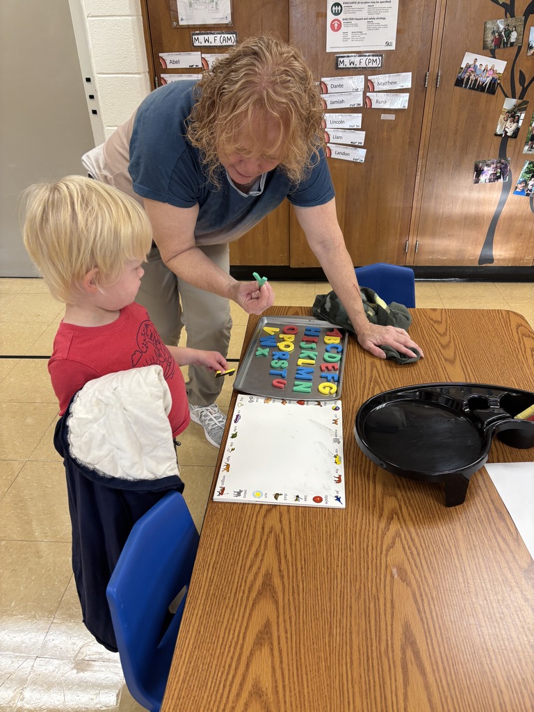 A teacher helps a young student practice letters with an alphabet learning board at a classroom table.