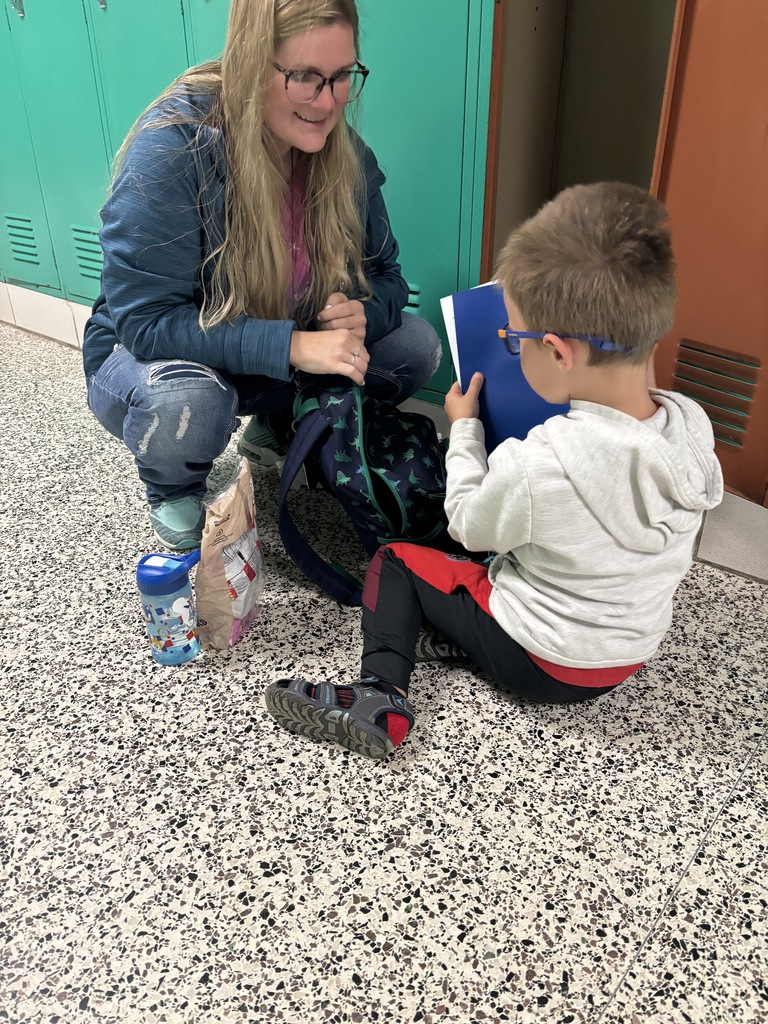 A woman crouches by hallway lockers to help a young student organize papers in a folder near his backpack.
