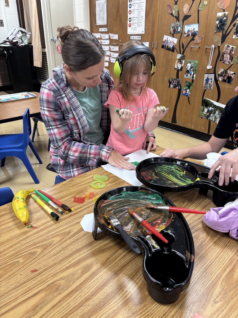 An adult assists a young student wearing noise-canceling headphones with a finger painting activity at a table.