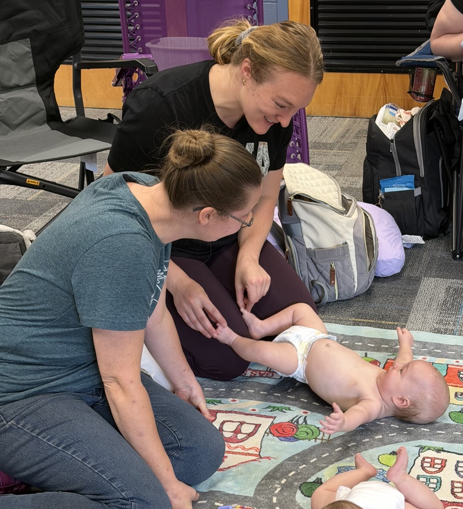Two adults smile while interacting with a baby lying on a play mat during an early childhood or family activity.