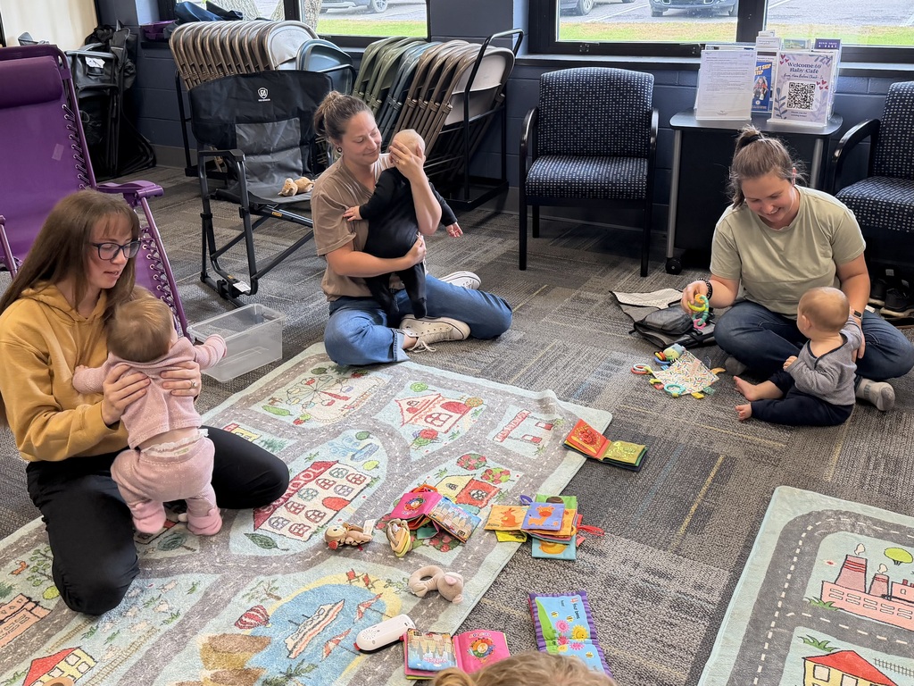 Three adults and babies play together with toys on a colorful mat during a Baby Cafe session.