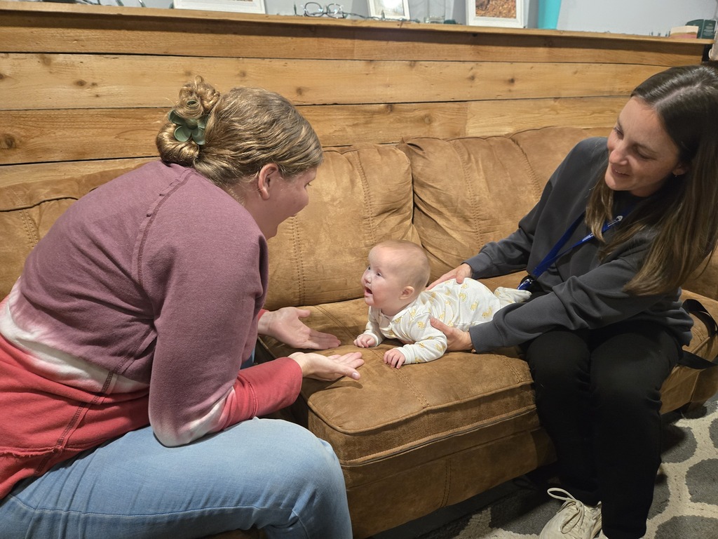 A baby lies on a couch smiling as two adults interact with and support the child during a home visit.