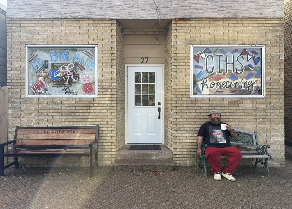 The front of Jojo Studio's building. The widows are decorated for homecoming. Jojo is sitting on a bench smiling, holding a coffee mug.