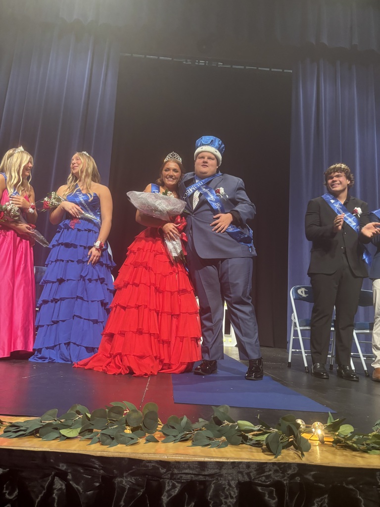 Cambridge-Isanti High School Homecoming royalty stand together on stage after being crowned, with the king wearing a blue crown and the queen holding flowers.