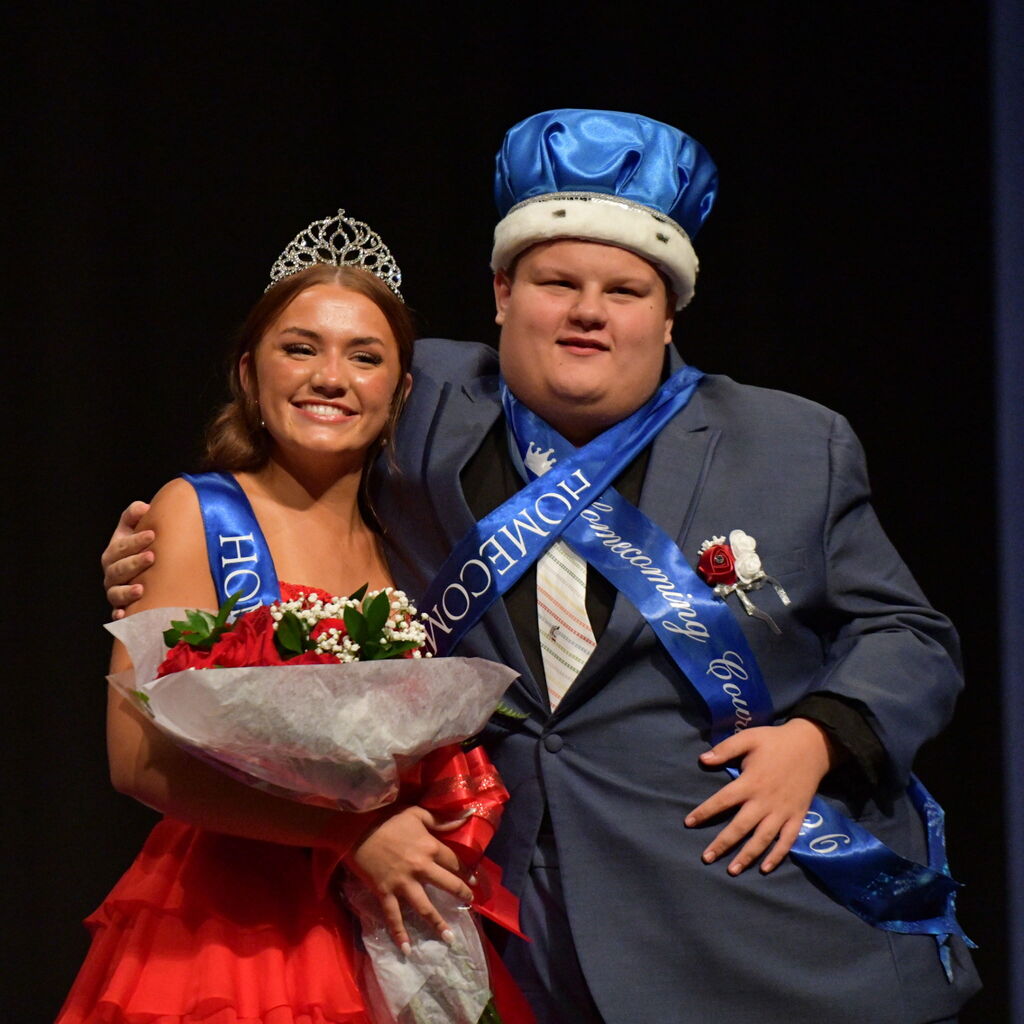 Two smiling students wear sashes and crowns while posing together with a bouquet of red roses during homecoming.