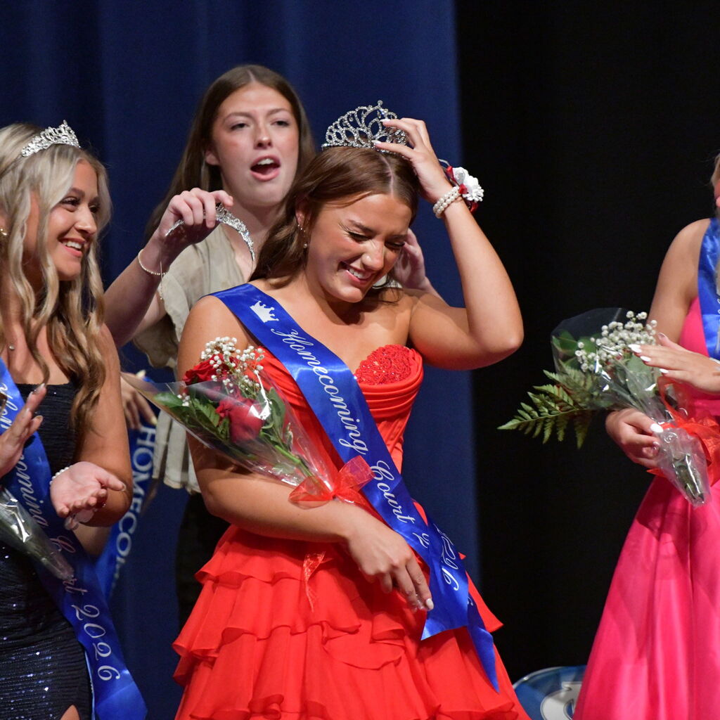 A student reacts with emotion as another student places a tiara on her head during a homecoming ceremony.