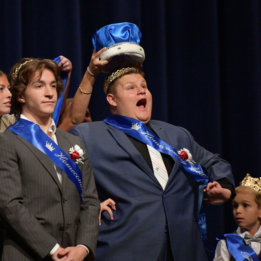 A student reacts with joy and excitement as a blue crown is placed on his head during a school homecoming ceremony.