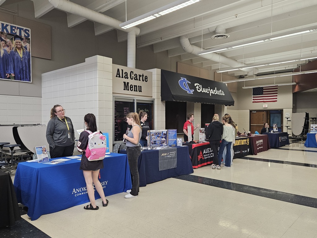 Students speak with representatives at informational tables set up in a school cafeteria during a college fair.