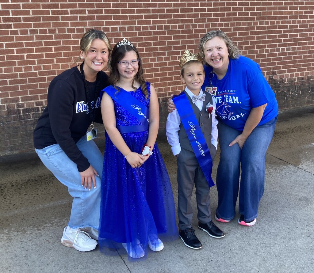 Two adults and two children standing together outdoors against a brick wall. A young girl in a blue sparkly gown and tiara with a sash, and a young boy wearing a crown, bowtie, vest, and sash, stand between two smiling women crouching on either side.