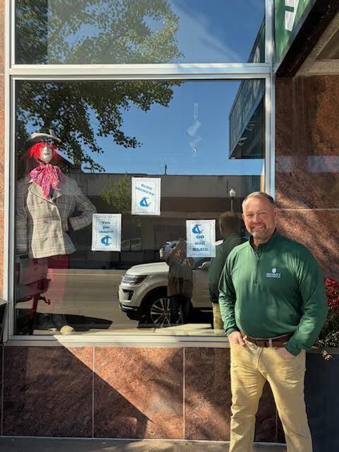 A smiling man stands outside a storefront displaying school spirit signs and a mannequin in a jacket.