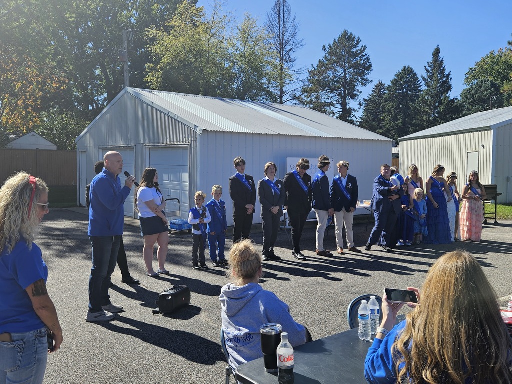The homecoming court is standing in a row. There is a man speaking into a microphone. It is a bright and sunny day.