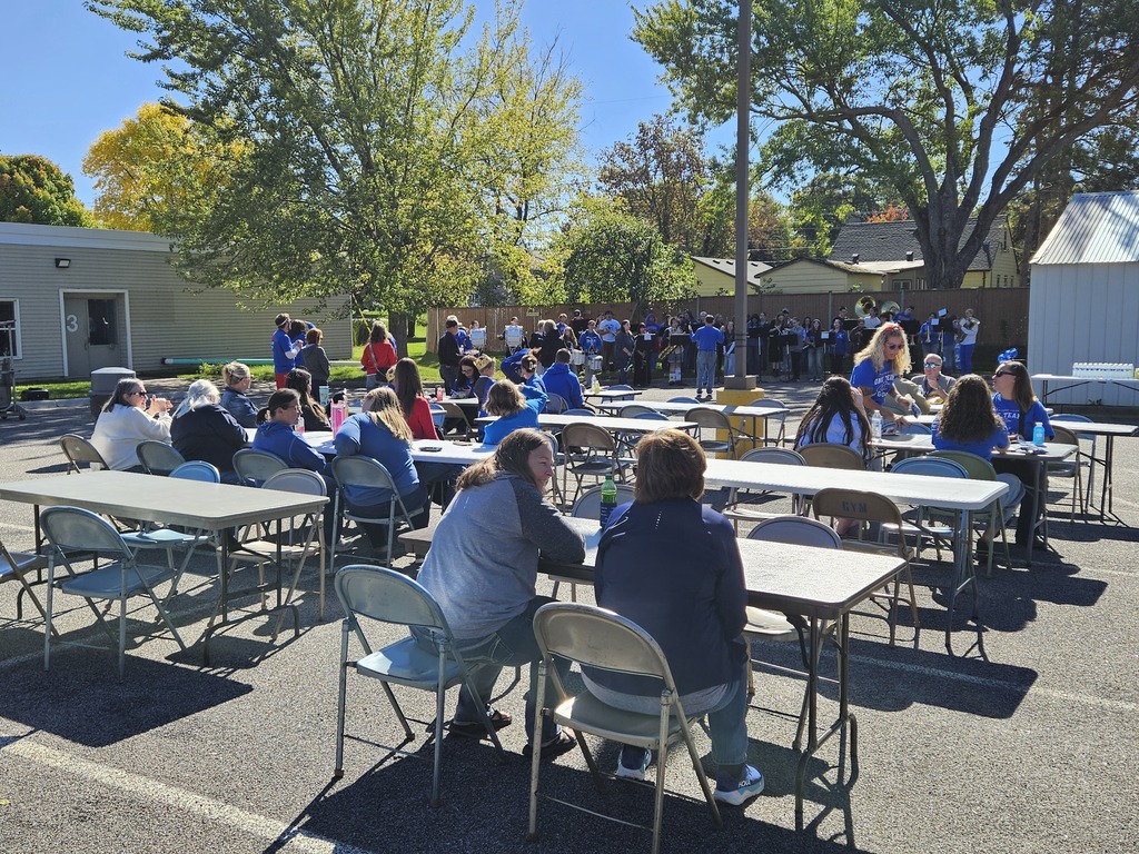 A group of people are sitting at tables outside in a parking lot. They are happy and socializing. It is a bright sunny day.