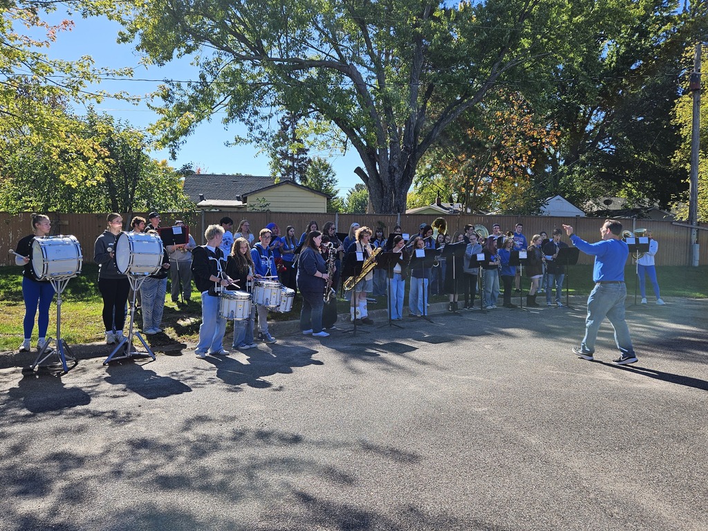 Marching band preforming for an audience on a bright sunny day. They are in a parking lot and a teacher is conducting.