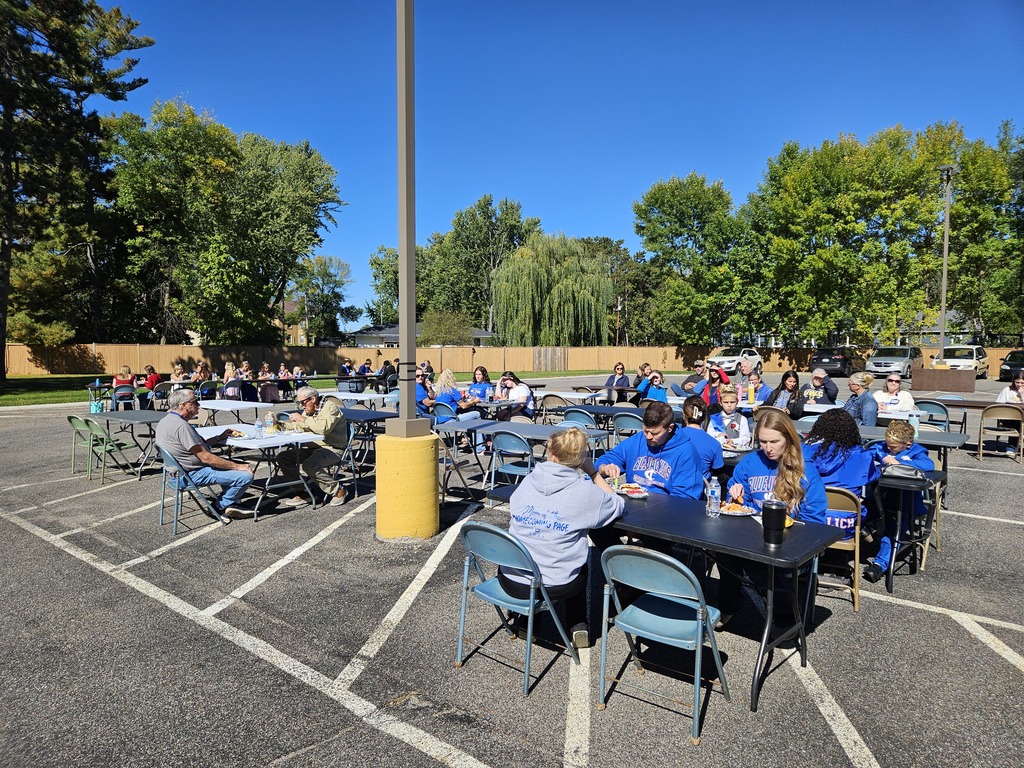 A group of people enjoying a picnic style lunch while sitting together at tables set up outside on a sunny day. 