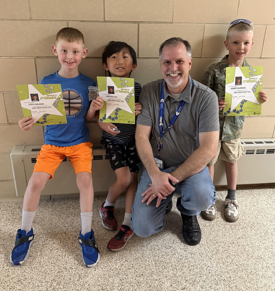 A smiling male school staff member kneels on the floor alongside three young boys holding green "Certificate of Attendance" star awards. The boy on the left wears a blue t-shirt with a basketball graphic and orange shorts; the boy in the middle wears a striped shirt; and the boy on the right wears a camo print shirt. They are posing together in front of a light brick wall inside a school hallway.