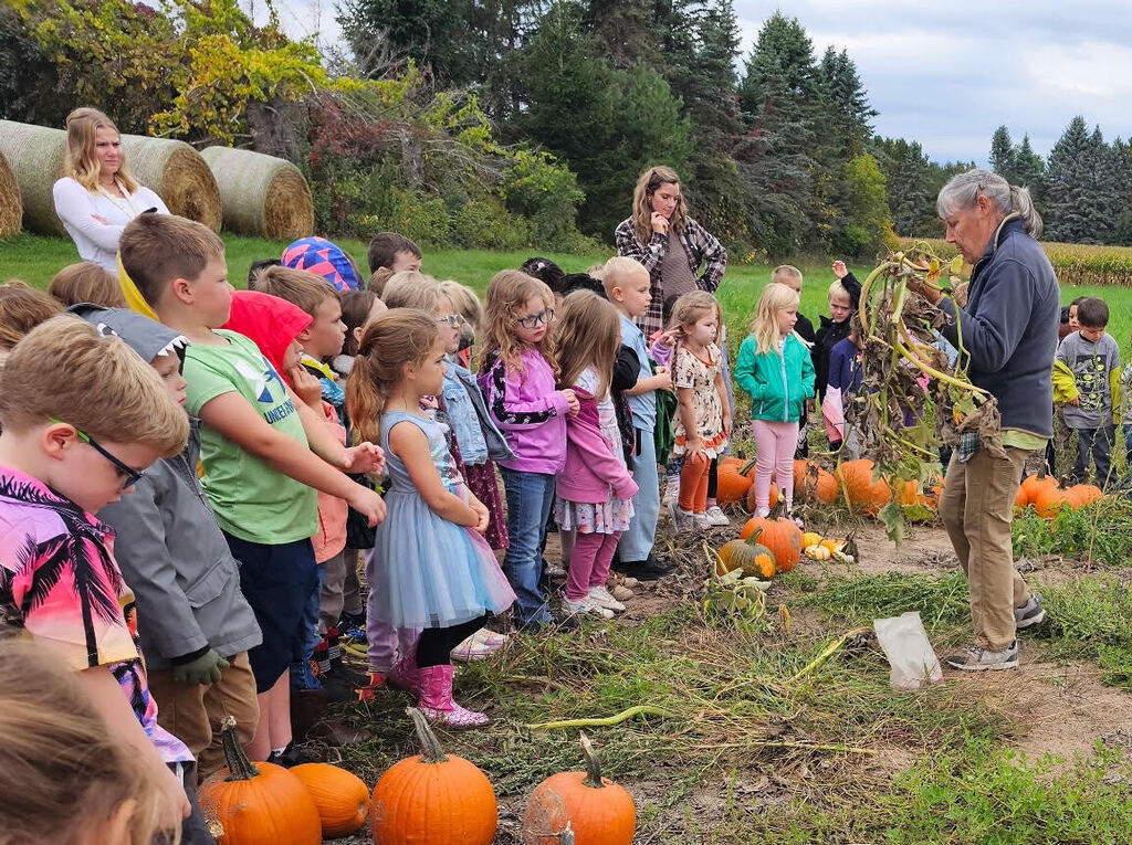 A group of young elementary students gathered in a pumpkin patch, gathered around an instructor holding up a dried pumpkin vine to teach them about how pumpkins grow.