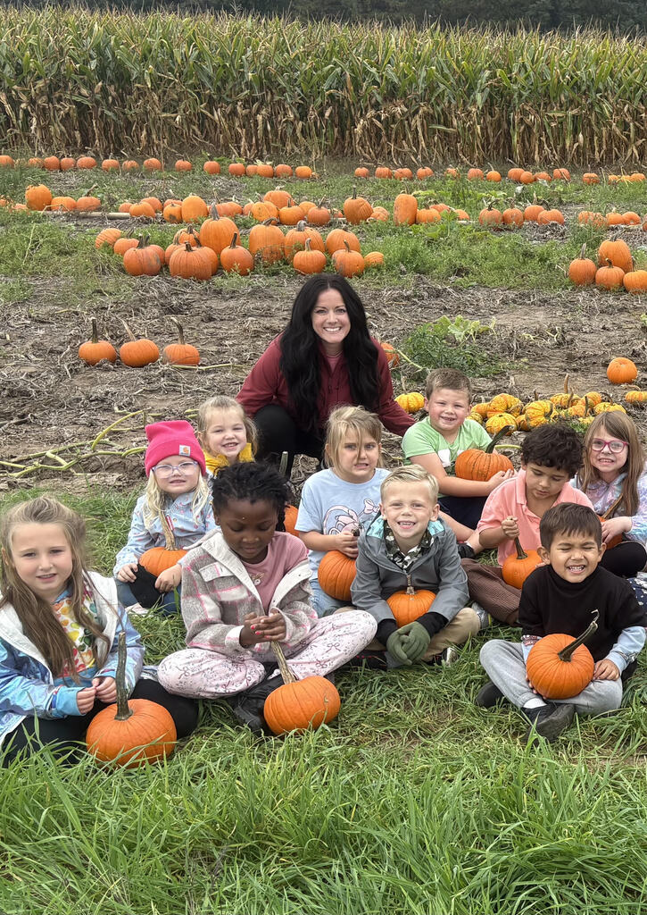 A teacher sitting in a pumpkin field surrounded by smiling kindergarten students, each holding or sitting near their chosen pumpkin.