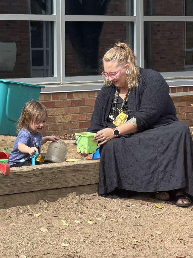 teacher sitting on the edge of a sandbox, and a student in the sandbox playing with sand toys
