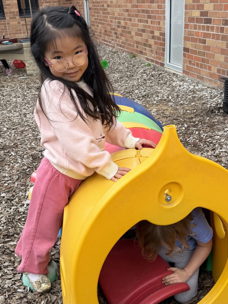 Two students playing on playground equipment, with one of them smiling wearing pink outfit and glasses and the other in a tunnel