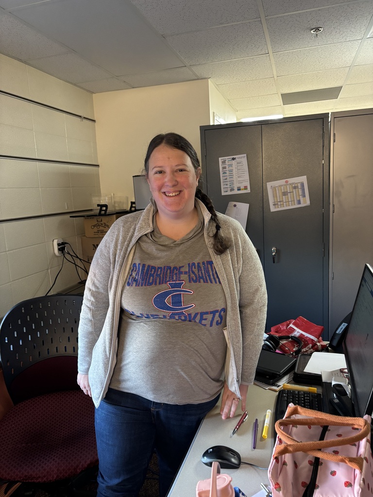 Teacher wearing a grey Bluejacket shirt smiles at camera in office area.