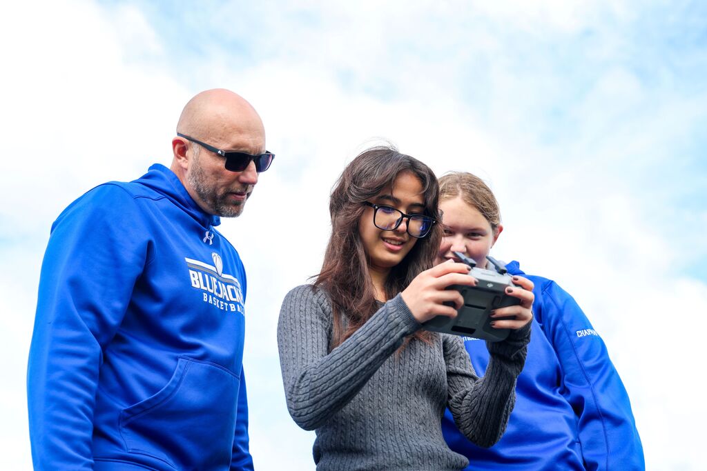 A teacher wearing a Bluejacket Basketball sweatshirt guides a student as she holds a drone remote controller outdoors.