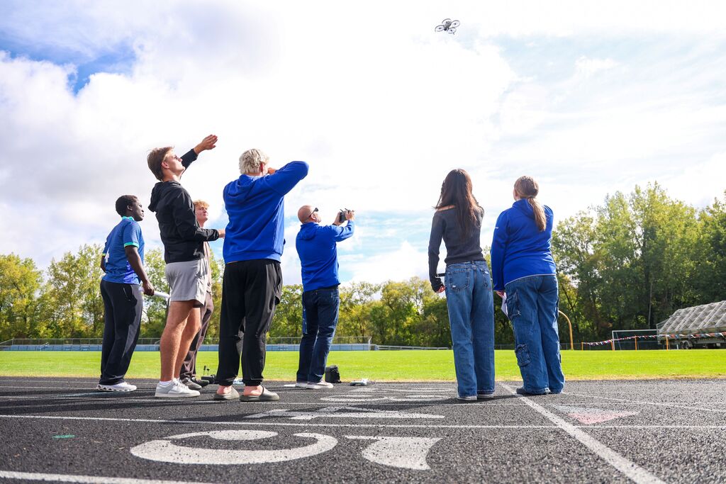 Students and staff gather on a running track and look up as a small drone flies overhead.