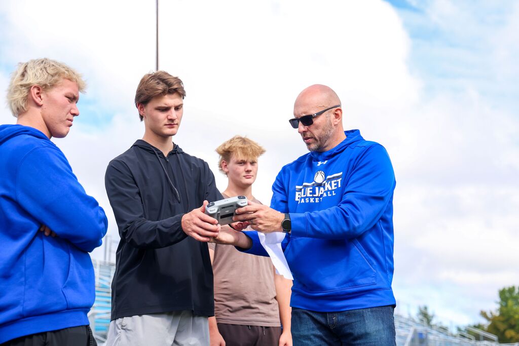 A teacher wearing a Bluejacket Basketball sweatshirt shows three students how to operate a drone controller outside.
